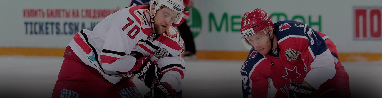 На изображении два хоккеиста в красной и белой форме борются за шайбу на льду. Видна часть рекламного баннера и логотип на бортике. Игра происходит в крытой ледовой арене