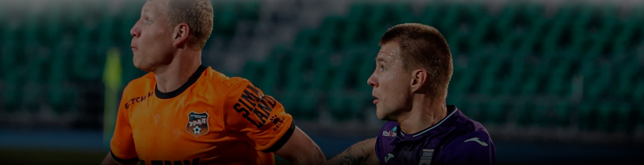 Два футболиста в оранжевой и фиолетовой форме смотрят в сторону, возможно, следя за мячом. На заднем плане размытые зеленые трибуны