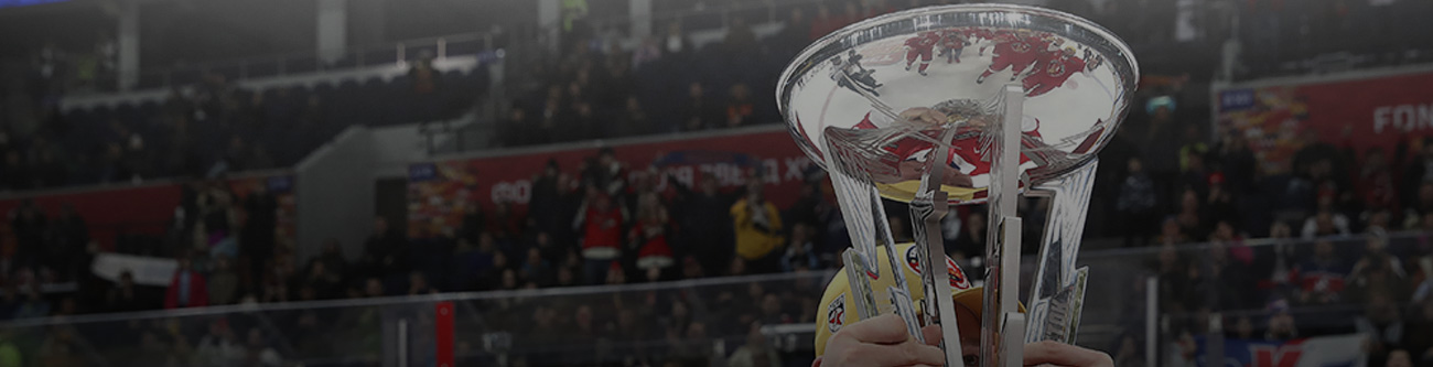 Спортсмен в яркой форме держит большой серебристый кубок. На заднем плане видны трибуны с болельщиками. Кубок блестит на свету, привлекая внимание