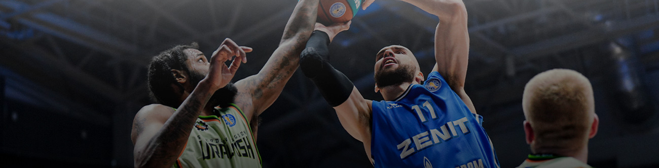 На изображении запечатлен динамичный момент баскетбольного матча. Два игрока борются за мяч в прыжке. Один из них в синей спортивной форме, другой — в зеленой. Фоном служит крытая спортивная арена